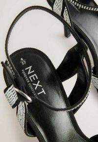 Sandales à talons noires avec une finition en cuir texturé, dotées d'une bride ornée d'argent et d'une semelle intérieure noire rembourrée portant le nom de la marque.