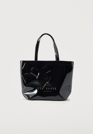 Sac fourre-tout noir brillant Ted Baker avec un grand nœud décoratif et un logo doré, deux poignées, sur un fond clair uni.