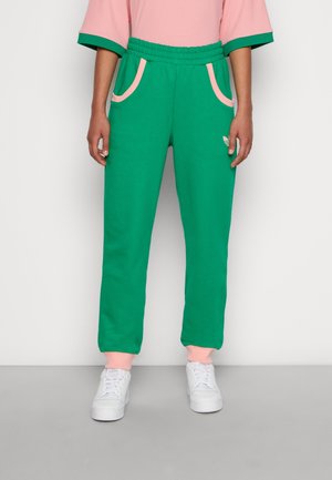 Persona che indossa pantaloni verdi con rifiniture rosa sulle tasche e sui polsini, abbinati a un top rosa e scarpe da ginnastica bianche, in piedi davanti a uno sfondo semplice.