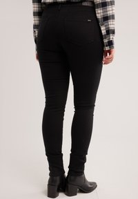 Inimene, kes kannab musta kitsast teksapükse, musti pahkluu-saapaid ja halli ruudulist särki, seisab heleda ühetaolise tausta ees.