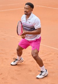 Tennis player in a white and gray striped polo with pink shorts, holding a pink tennis racket, on a red clay court. White athletic shoes and socks.