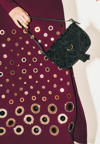 Borsa a tracolla nera con perline, con una parte superiore curva e un dettaglio ad anello dorato, tenuta contro un vestito bordeaux ornato con varie paillettes metalliche.