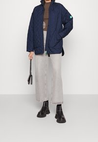 Mujer vistiendo una chaqueta acolchada azul marino, blusa marrón, pantalones anchos de mezclilla gris claro, botas negras gruesas y sosteniendo un pequeño bolso negro.