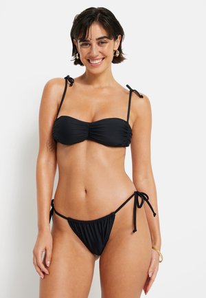Čierny bikini s bandeau vrcholom, ktorý má riasenie a nastaviteľné viazanie, v kombinácii s nízko strihaným spodným dielom so šnúrkami po stranách. Hladká textúra.