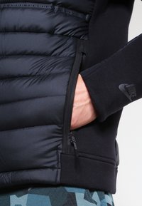 Černá péřová bunda s žebrovanou tkaninou, vybavená boční kapsou na zip. Design zdobí logo Nike na rukávu.