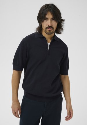 Mand med mellemlangs mørkt hår og skæg iført en mørk tekstureret kortærmet lynlåstrøje og mørke bukser, stående foran en ensfarvet baggrund.