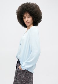 Hele sinine pluus lahtise lõikega, ümar kaelus ja pikkade varrukatega, kombineeritud mustrilise seelikuga, millel on halli ja burgundi toone.