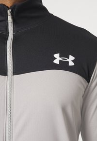 Gros plan d'une veste zippée bicolore gris et noir avec un col haut et un logo blanc Under Armour sur la poitrine.