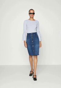 Camisola cinza-claro de manga longa com textura canelada; saia lápis de denim azul de cintura alta com fecho de botão na frente; sapatos pretos de salto bloco.