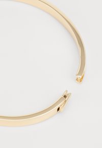 Gold metal bangle with a sleek, curved design and a shiny finish. Features a subtle opening mechanism near the ends for easy wear.