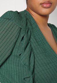 Gros plan sur l'épaule et le cou d'une personne portant un chemisier vert foncé texturé avec des détails de manches à volants.