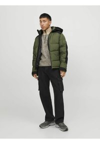 Giacca verde gonfiata con accenti neri, chiusura lampo frontale e cappuccio. Indossata sopra un maglione leggero e testurizzato e abbinata a pantaloni cargo neri. Sneakers grigie.