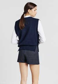 Un pull tricoté sans manches bleu marine porté sur une chemise à col blanc, associé à un short à rayures grises foncées avec une coupe légèrement ample.