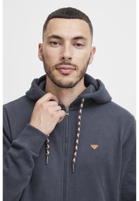 Mornarska zip-up dukserica s kapuljačom na vezanje, teksturirana tkanina i mali bež logotip. Ima pletene vezice s bež, crnim i bijelim detaljima.