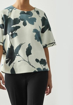 Femme portant un haut blanc à manches courtes avec un grand imprimé floral bleu foncé, associé à un pantalon noir, debout devant un fond uni.
