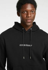 Felpa nera con cappuccio in tessuto morbido, dotata di tasca frontale, coulisse e logo bianco "GUESS JEANS" sulla parte anteriore del petto.