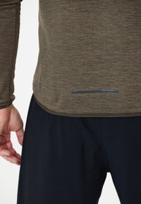 Langärmliges Shirt aus strukturiertem olivgrünem Stoff mit abgerundetem Saum und einem reflektierenden Akzent an der Unterseite, kombiniert mit dunkelblauen Shorts.