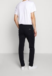 Zwarte broeken met een elastische tailleband, voorzien van een klein metalen logo op de achterkant. Gecombineerd met een eenvoudig wit T-shirt en witte sneakers.
