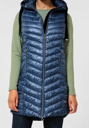 Close-up van een persoon die een marineblauwe doorgestikte puffer-vest met capuchon en ritszakken draagt, over een groen shirt met lange mouwen.