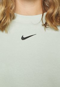 Svetle zelená mikina vyrobená z měkké látky, s černým vyšitým logem Nike uprostřed. Žebrovaný výstřih dodává detail.
