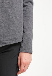 Grå långärmad topp gjord av mjukt tyg; har en slät yta och en något rundad nederkant. Handen vilar på svarta byxor.