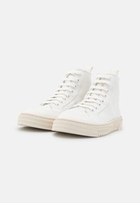 Paire de baskets blanches montantes en toile avec semelles en caoutchouc beige et lacets, placées côte à côte sur un fond blanc.