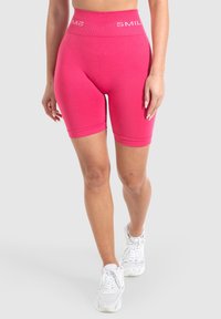 Roze stretchshorts met een hoge tailleband met de tekst "SMIL". Gladde textuur, aansluitend ontwerp en lengte tot halverwege de dij. Gepaard met witte sneakers.
