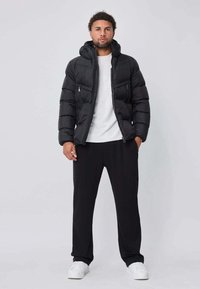 Doudoune noire avec capuche, dotée de poches zippées. Portée sur une chemise blanche, associée à un pantalon noir et des baskets blanches.