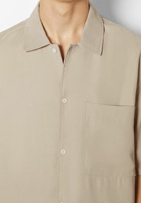 Short-sleeve beige shirt made of smooth fabric, featuring a pointed collar, front pocket, and button closure. Minimalist design with subtle texture.