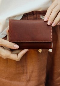 Brown leather wallet with stitched edges, compact rectangular shape, and a smooth texture. Held in a hand against a rust-colored background.