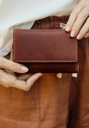 Braune Lederbörse mit genähten Kanten, kompaktem rechteckigen Format und glatter Textur. In einer Hand vor einem rostfarbenen Hintergrund gehalten.