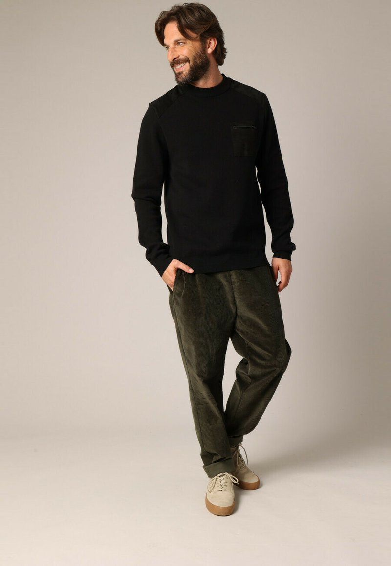 Zwarte longsleeve sweatshirt met een textuur op het schouderpaneel en een borstzak, gecombineerd met olijfgroene corduroy broek en lichtbeige sneakers.