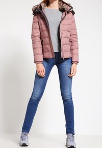 Różowa puchowa kurtka z futrzanym kapturem, kieszeniami na zamek oraz elastycznymi mankietami, połączona z szarym topem i niebieskimi dżinsami skinny, dopełniona szarymi sneakersami.