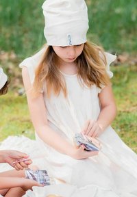 Tyttö, jolla on päällään vaalean, ilmanhihoja oleva mekko ja vaaleanbeige pipo, pitää värikkäitä pelikortteja käsissään istuessaan nurmikkoalueella, luonnon ympäröimänä.