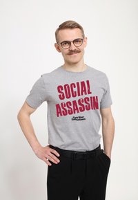 Camiseta de algodón gris con texto rojo en negrita "SOCIAL ASSASSIN" y el logo de "Curb Your Enthusiasm" debajo, combinada con pantalones negros y cinturón.