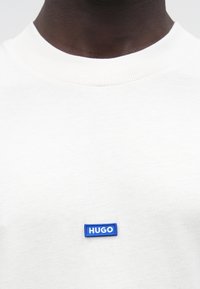 Κοντινό πλάνο ενός ατόμου που φοράει λευκό μπλουζάκι με στρογγυλή λαιμόκοψη και έναν μικρό μπλε ορθογώνιο λεκέ με την επιγραφή "HUGO" στο στήθος.