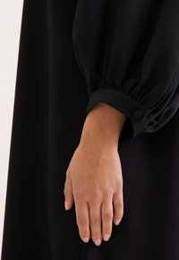 Schwarze Bluse mit langen, gepufften Ärmeln und einem geknöpften Bündchen. Glatte Stoffstruktur und taillierte Handgelenkdetails.