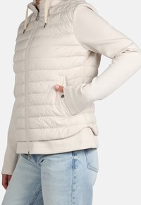 Chaleco tipo puffer en beige claro con costuras horizontales, cierre de cremallera y bolsillos laterales. Puños de tela suave en un tono que combina. Ajuste casual.