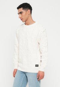 Man in een gestructureerde witte kabeltrui en lichtblauwe spijkerbroek, kijkend naar de zijkant tegen een effen witte achtergrond.
