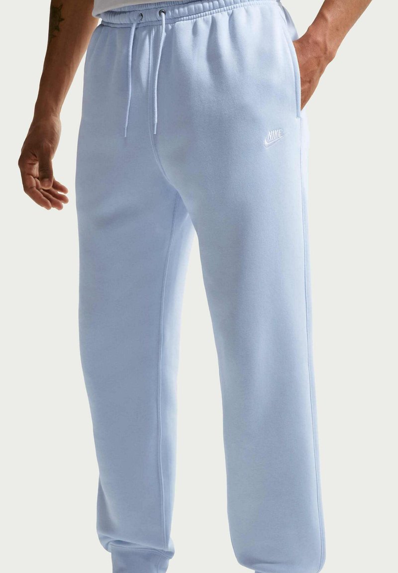 Persona che indossa pantaloni della tuta Nike azzurri con fascia elastica e coulisse, una mano in tasca, in piedi contro uno sfondo neutro.
