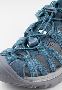 Blue water sandal with textured fabric straps, grey rubber sole, elastic lacing system, and cut-out design for breathability.