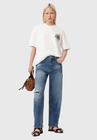 Wit T-shirt met bloemrijke borduursels op de borst, blauwe hoge taille jeans met versleten details, bruine suède tas en sandalen met print.