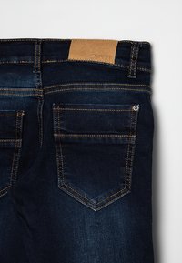 Jean en denim bleu foncé, vue de dos, avec des poches à double surpiqûre et un patch en cuir marron de la marque Esprit sur la ceinture.