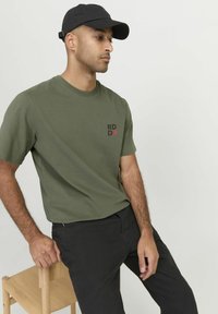 Camiseta de algodón color verde oliva con mangas cortas, que presenta un pequeño logo negro y rojo en el lado izquierdo del pecho. Combinada con pantalones negros.
