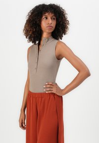 Woman with curly hair wearing a sleeveless ribbed beige top with buttons and high-waisted loose rust-colored pants, standing with hand on hip.