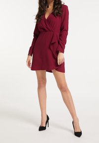 Bordeaux-Wickelkleid mit langen Ärmeln, einem gerüschten Saum und einem tiefen V-Ausschnitt. Kombiniert mit schwarzen Stiletto-Heels. Glatte Textur.