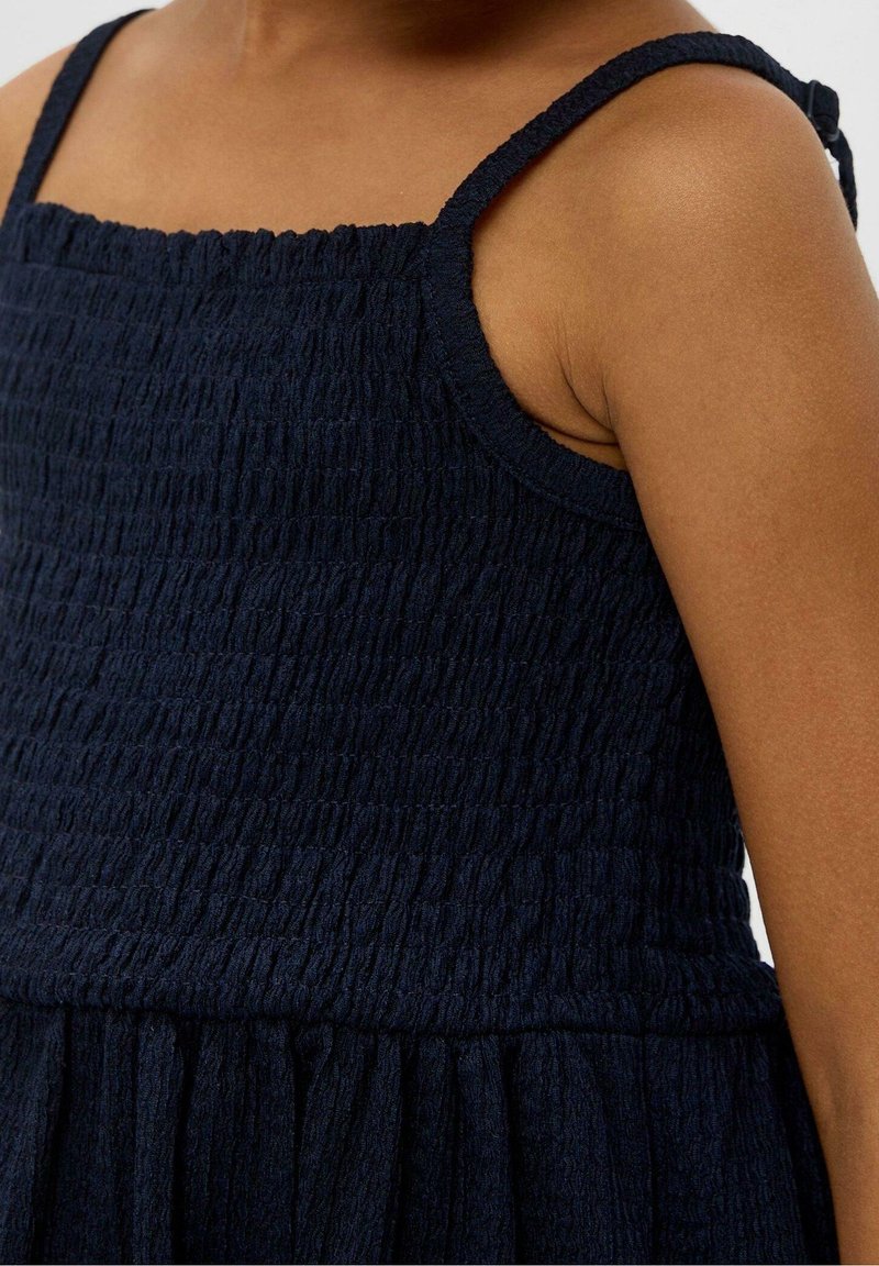 Enfant portant une robe sans manches texturée bleu marine avec de fines bretelles, montrant le torse supérieur et le bras.