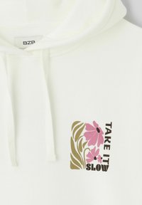 Sweat à capuche blanc avec un motif floral rose et des feuilles vertes à côté du texte "PRENDS TON TEMPS" sur la poitrine.