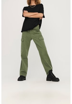 Persona de pie con los brazos cruzados, llevando una camiseta negra holgada, pantalones cargo verde oliva y botas negras con plataforma y cordones.