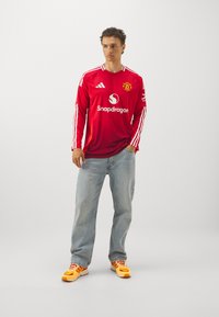 Raudona ilgomis rankovėmis džersio maikė su baltais dryžiais, su logotipu ir rėmėjo tekstu. Suderinta su šviesiai mėlynomis džinsomis ir geltonais sportiniais bateliais.